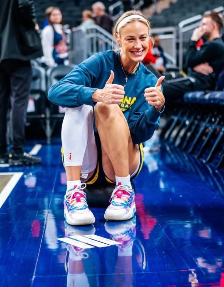 A smiling woman in athletic wear, possibly a basketball player, crouches on a blue court, giving two thumbs up. She wears a blue sweatshirt with 'ANA BA' visible, white knee-high socks with a red exclamation mark, and colorful sneakers. Spectators are in the background.