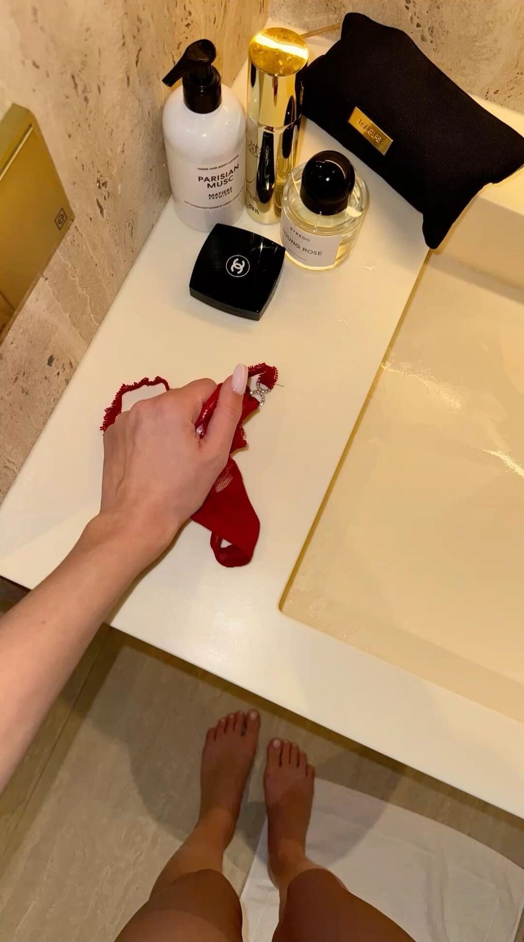 A bathroom counter with various vanity bottles and a black pouch. A hand grips a red cloth or fabric on the counter, with bare feet visible below.