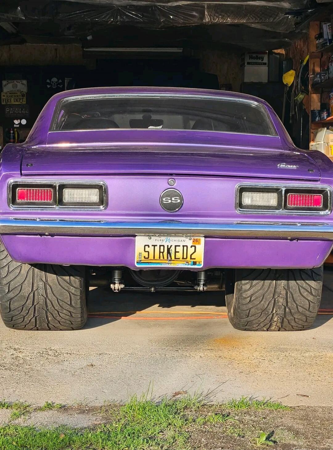 A purple classic car, possibly a Chevrolet Camaro, with wide rear tires, parked in a garage. The license plate reads 'STRKED2' from 'PURE MICHIGAN' and has '26' on it. The car features an 'SS' emblem. In the background, there are signs like 'Holley' and 'HARTFORD FIRE DEPT. PA-4444' with a skull and crossbones symbol.