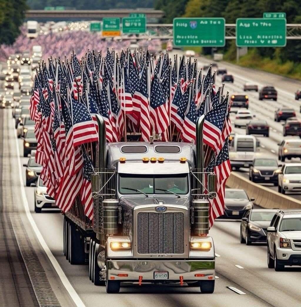 A large semi-truck, heavily decorated with numerous American flags, is driving on a multi-lane highway. Other vehicles are visible in traffic, and road signs can be seen in the background.