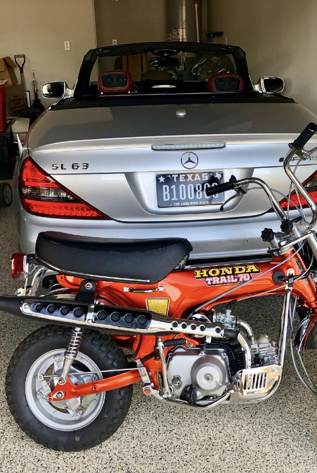 A silver Mercedes-Benz SL 63 convertible with a Texas license plate B1008C is parked in a garage behind an orange vintage Honda Trail 70 (CT70) motorcycle.