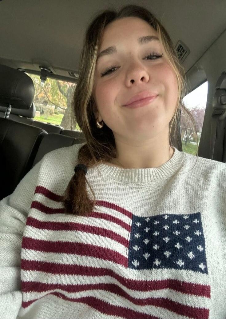 A young woman smiling in a car, wearing a sweater with an American flag design.