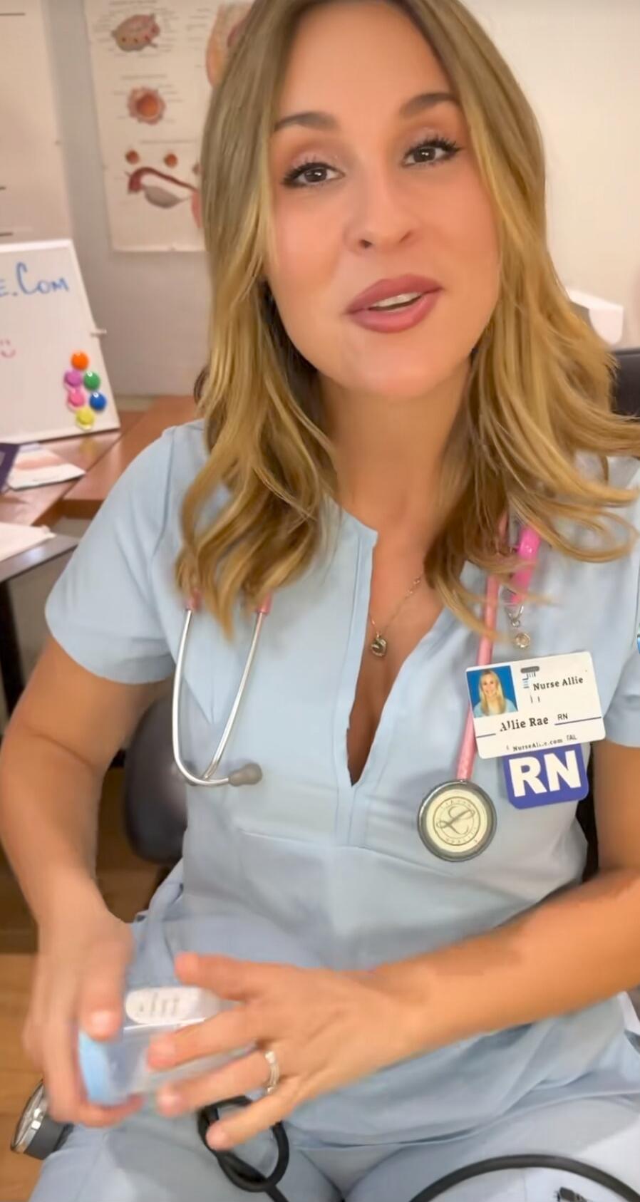 A nurse in light blue scrubs with a stethoscope around her neck, wearing an RN badge with a photo ID. She is in a medical office.