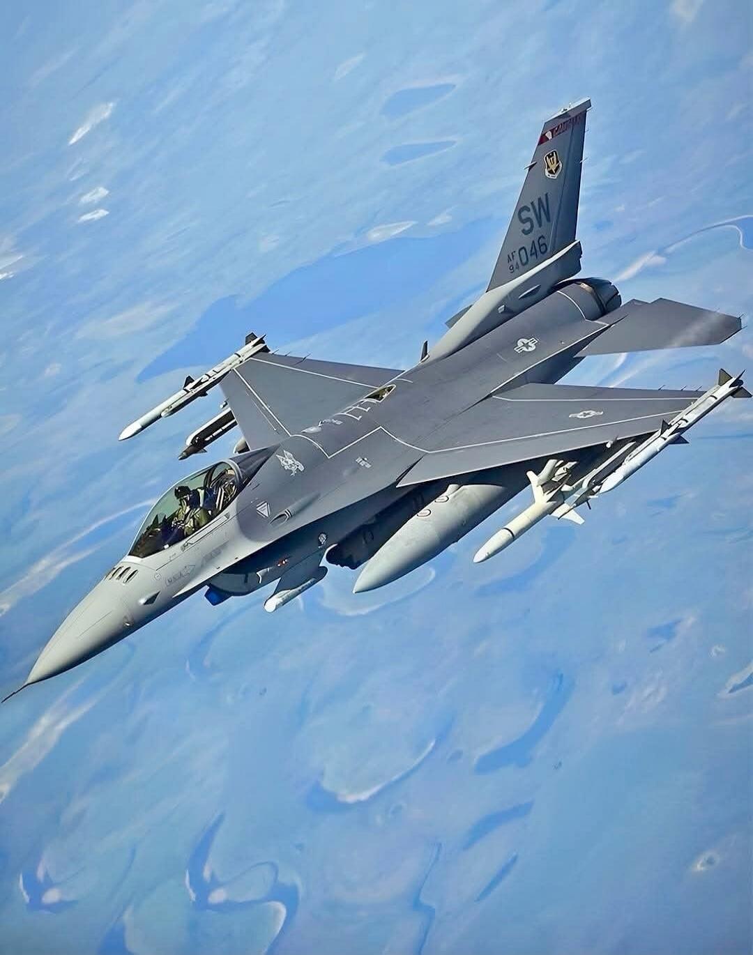 An F-16 Fighting Falcon fighter jet in flight, seen from above, against a blue sky with scattered clouds or land formations below. The aircraft is grey, armed with missiles and external fuel tanks, and has 'SW AF 94 046' marked on its tail fin.