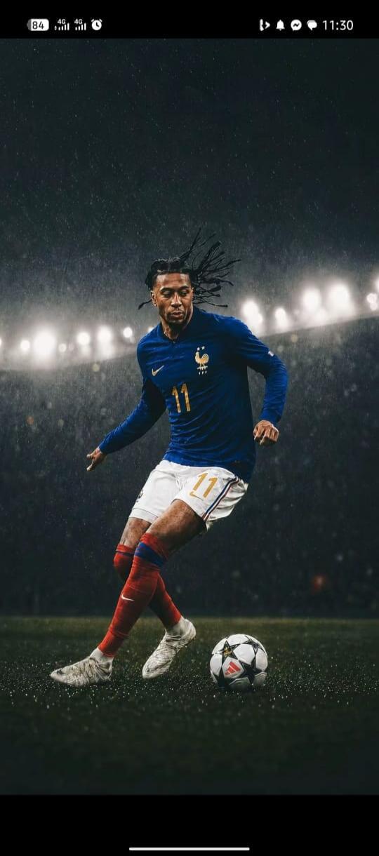 A male football player wearing a blue jersey with number 11, white shorts, and red socks is on a green field with a soccer ball. It appears to be raining, and stadium lights are visible in the background.