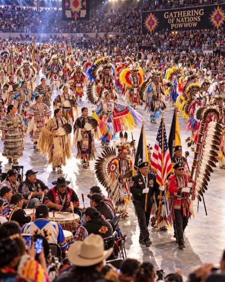GATHERING OF NATIONS POWWOW. A large group of people in traditional Native American regalia are dancing and participating in a powwow in a large arena. There are drummers playing, and some individuals are carrying flags, including the American flag. A large audience is visible in the stands.