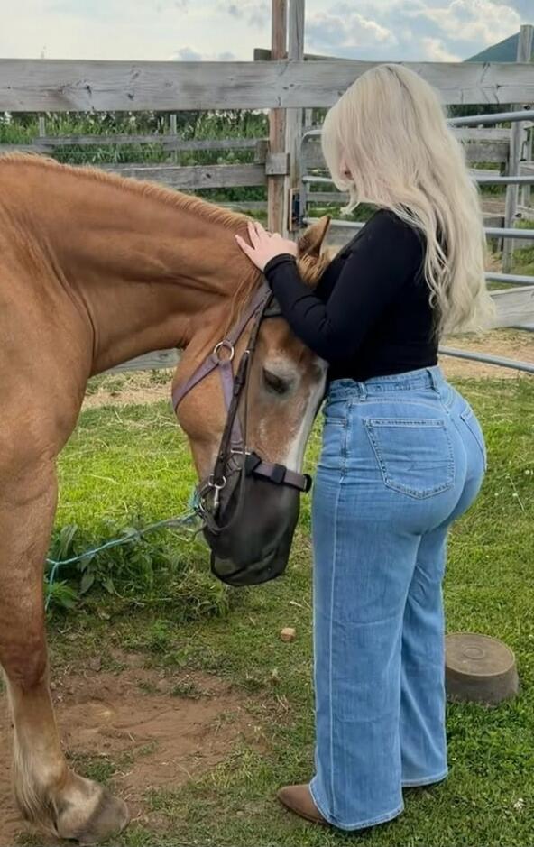 A woman with long blonde hair is gently petting a brown horse's neck. The horse is wearing a fly mask. They are outdoors in a grassy area with a wooden fence in the background.