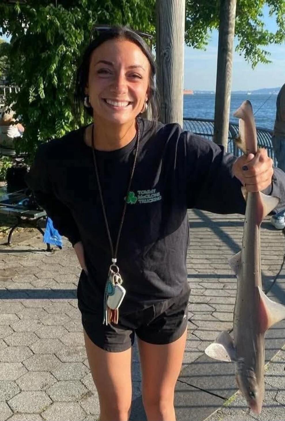 A young woman smiling and holding a small shark by its tail. Her black long-sleeve shirt has the text 'TOMMY MCCLONE TRUCKING' with a shamrock logo.