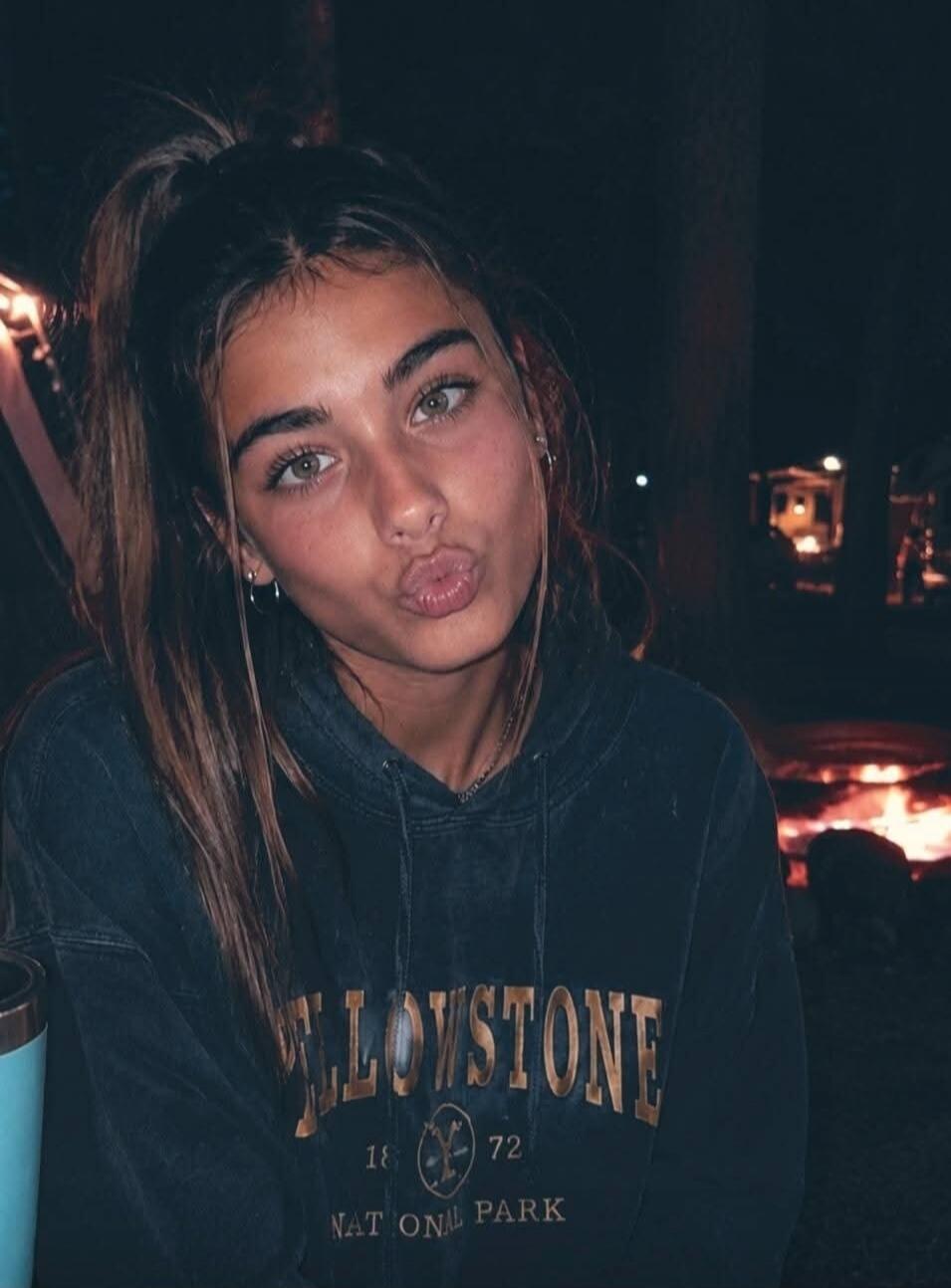 YELLOWSTONE 18 72 NATIONAL PARK. A young woman with long hair in a ponytail, wearing a Yellowstone National Park hoodie, makes a kissing face at the camera. A campfire is visible in the dark background.