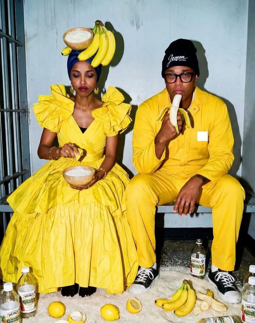 Two people wearing bright yellow outfits sit together. The woman has a hat made of bananas and a bowl in her lap; the man is eating a banana. There are bananas and bottles on the floor around them.