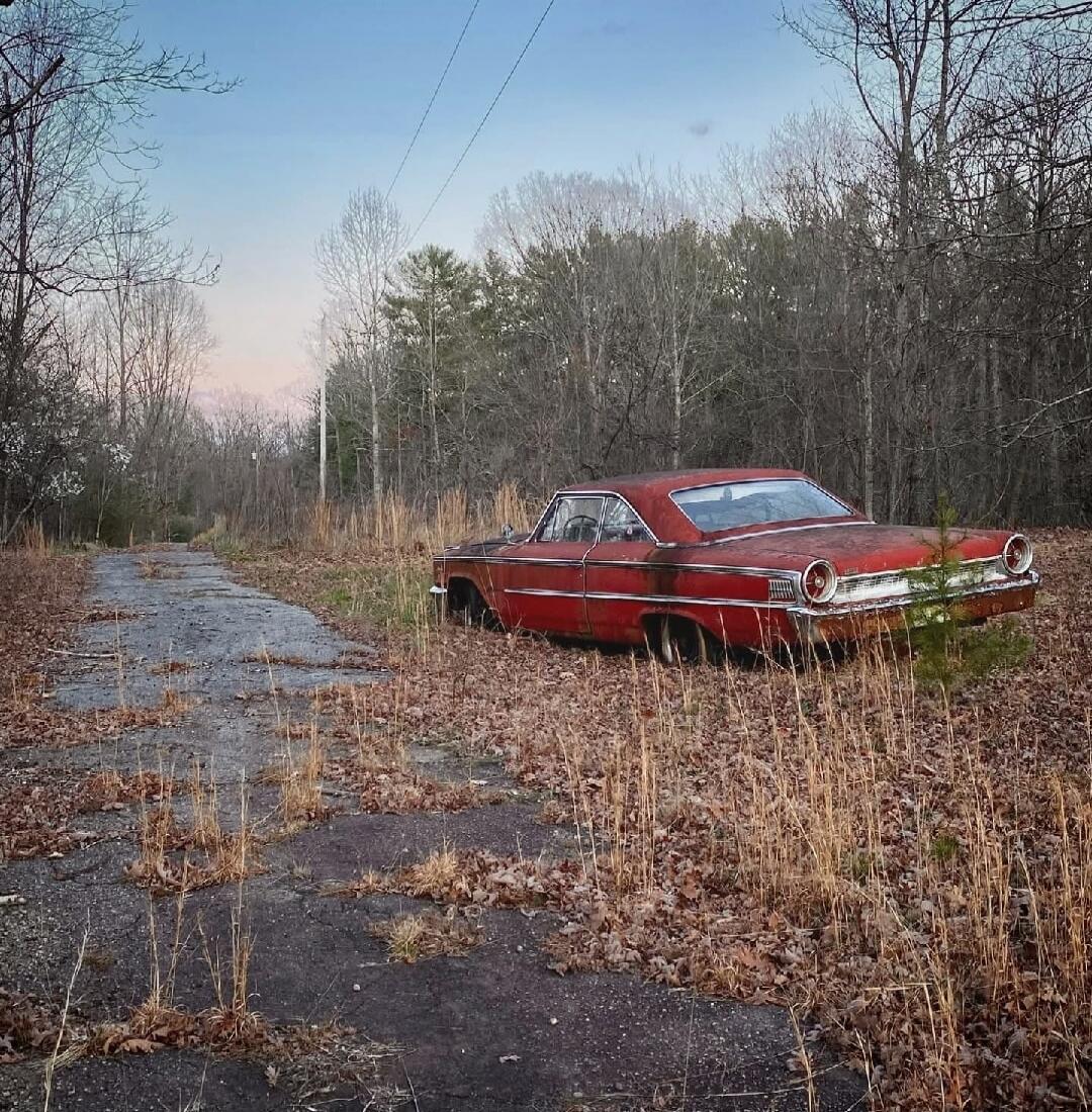 An old red car abandoned on a cracked, overgrown road in a rural/forest area.
