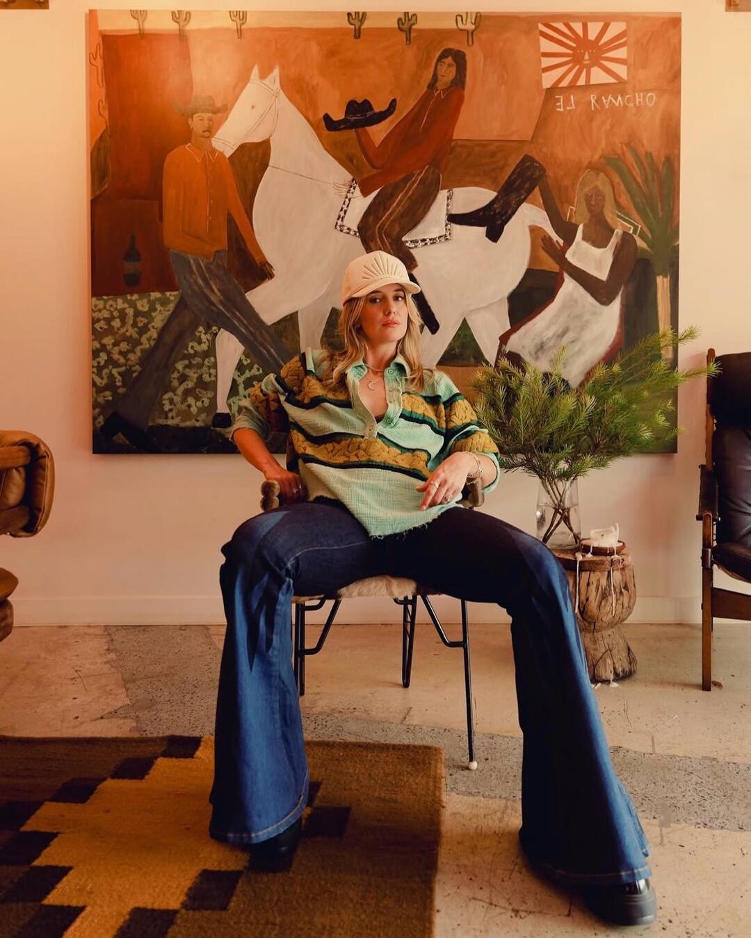 A woman wearing a striped shirt, wide-leg jeans, and a white cap sits in a chair in front of a large painting depicting horse riders.