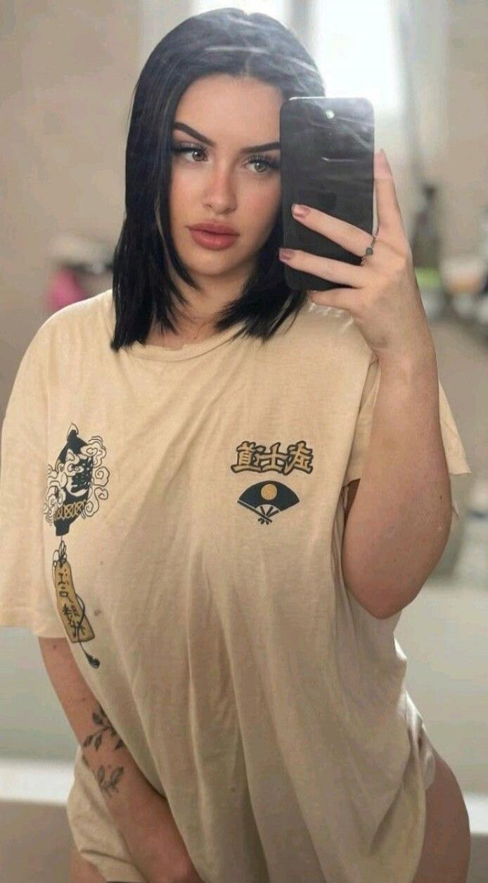A young woman takes a mirror selfie, holding a phone to her face. She wears an oversized beige t-shirt featuring Asian-inspired graphics, including a stylized demon face, a traditional fan, and some decorative characters. Her dark hair is styled around her face, and she has noticeable makeup. A tattoo is visible on her left forearm, and her bare ri
