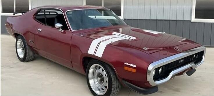 A classic maroon muscle car, a Plymouth GTX, with white racing stripes on the hood and side. The car features '440' and 'GTX' badges. It is parked on a concrete surface in front of a building.
