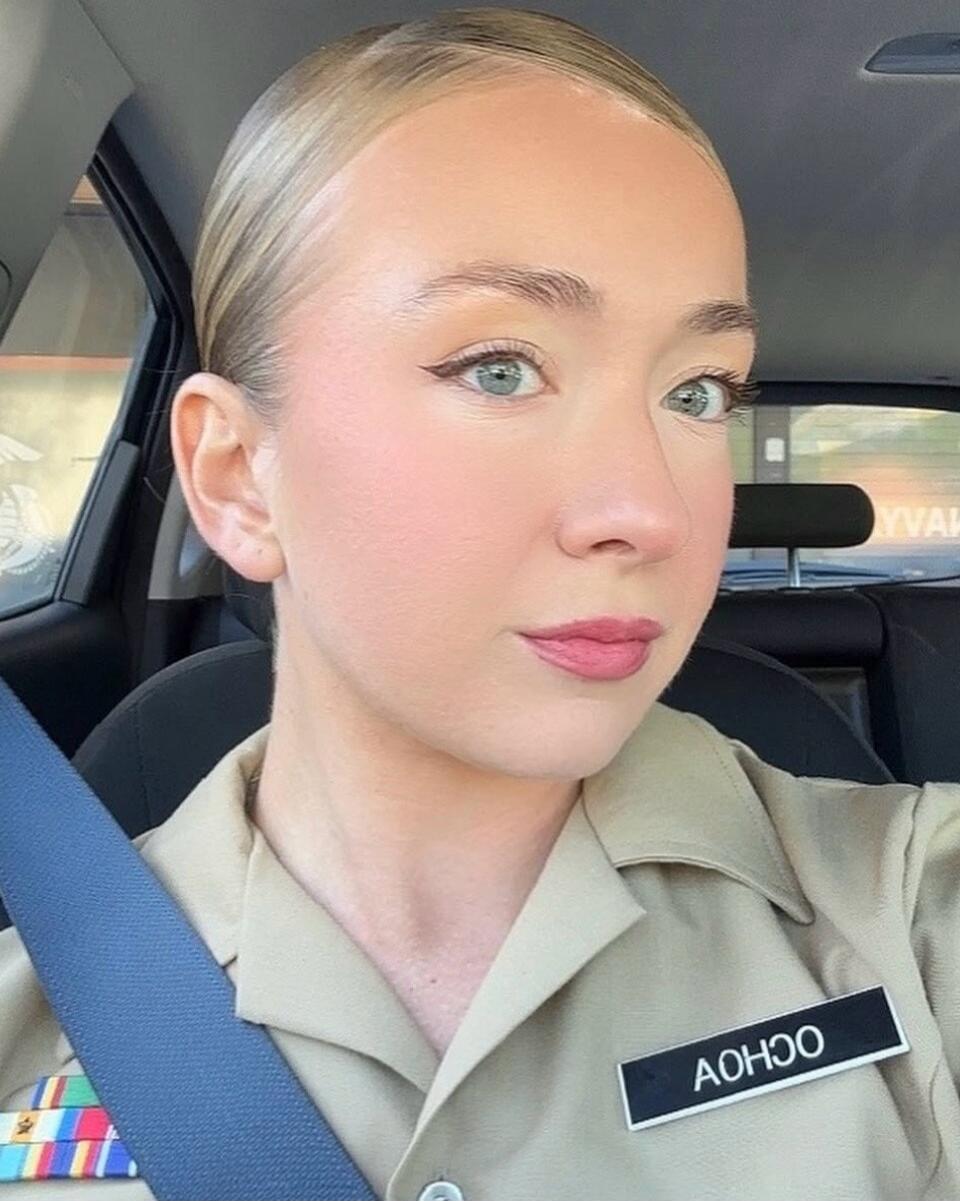 A woman in a uniform with a name tag that reads 'AOHOO' is taking a selfie in a car. She is wearing a seatbelt and has ribbons on her uniform.