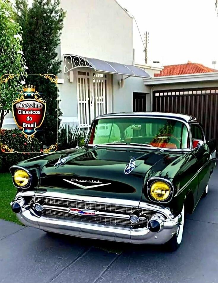 Green classic 1957 Chevrolet Bel Air parked in a driveway in front of a house. A decorative sign on the left reads “Magazine Classicos do Brasil.”