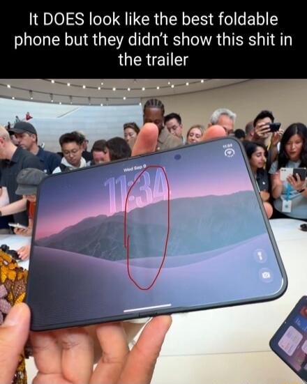 It DOES look like the best foldable phone but they didn't show this shit in the trailer. The image shows a person holding a foldable phone with a visible crease on the screen, highlighted by a red circle. The phone displays the time '11:34' and a mountain wallpaper. People are visible in the background, suggesting a tech event or product launch.