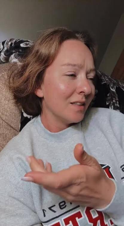 A woman with a thoughtful or slightly exasperated expression, making a gesture with her hand, palm up. Partial text 're' and 'T Я' visible on her grey sweatshirt.