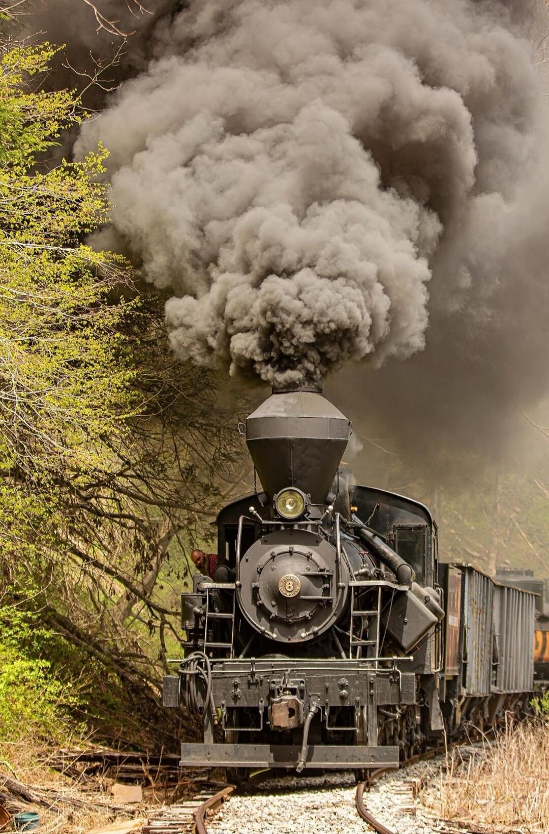 A steam locomotive emits a large plume of dark smoke as it travels on a track through a wooded area.