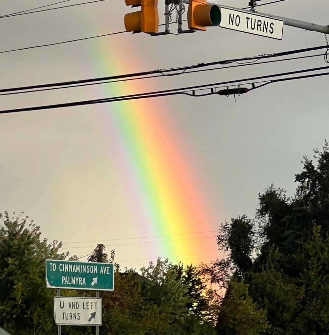A vibrant rainbow arches across a cloudy sky, with traffic lights and street signs in the foreground. The signs read 'NO TURNS', 'TO CINNAMINSON AVE PALMYRA', and 'U AND LEFT TURNS'.