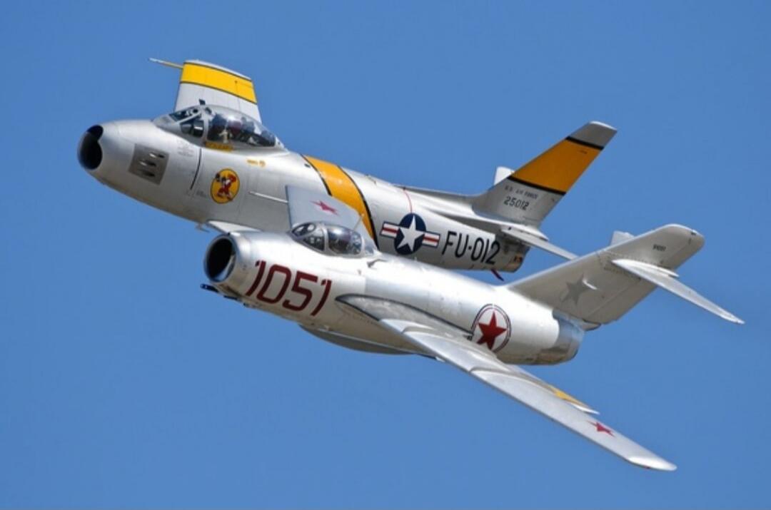 Two vintage jet fighter aircraft, a North American F-86 Sabre (marked 'U.S. AIR FORCE 25012 FU-012') and a Mikoyan-Gurevich MiG-15 (marked '1051' with a red star), fly in close formation against a clear blue sky. The F-86 is above and slightly behind the MiG-15.