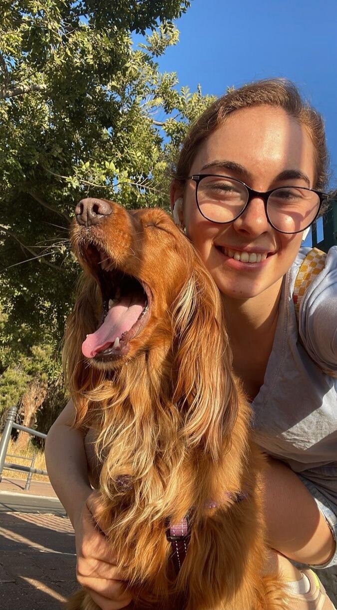 A young woman with glasses is taking a selfie with a brown long-haired dog outdoors on a sunny day. The dog is yawning widely, showing its tongue and teeth, while the woman smiles.