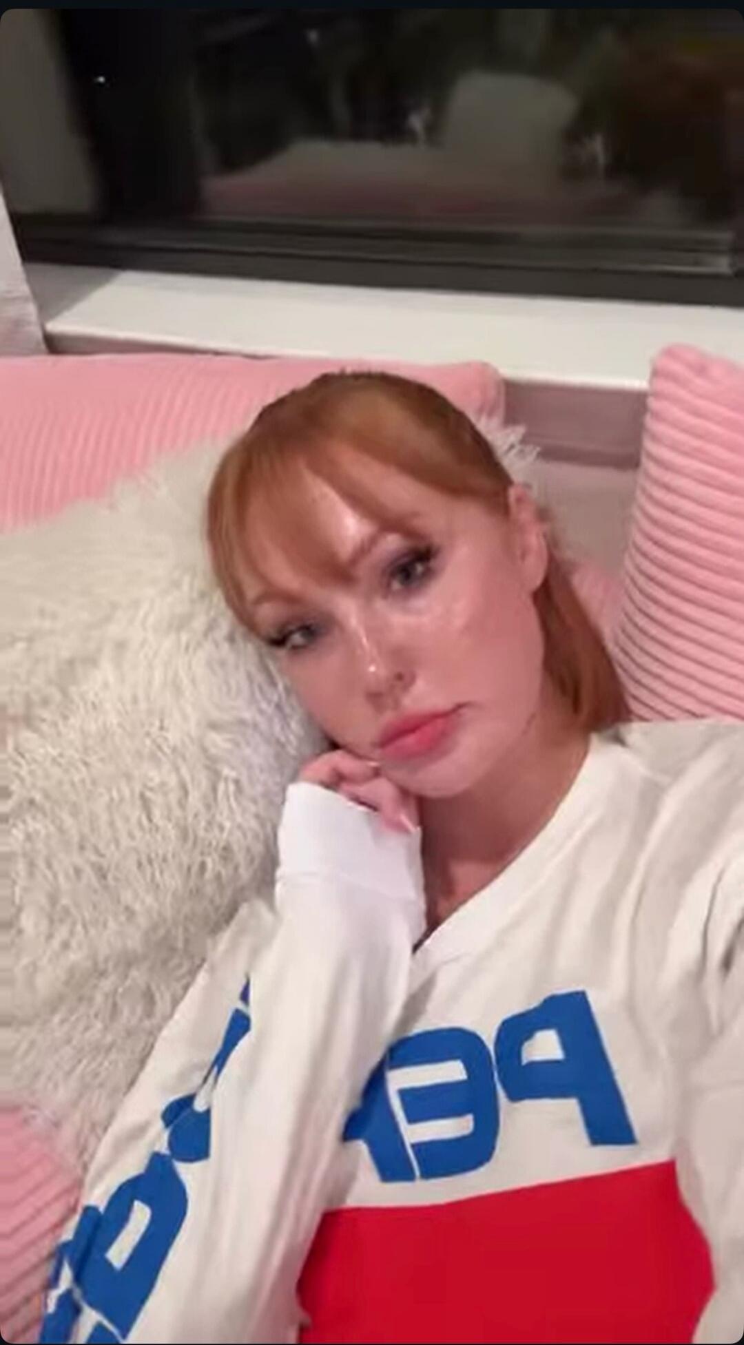 A woman with red hair is lying on a bed or sofa, resting her head on a white fluffy pillow. She is wearing a white long-sleeve shirt with a red and blue design, which appears to be a mirrored 'PEPSI' logo. Pink pillows are visible in the background. She has a neutral or slightly pensive expression.