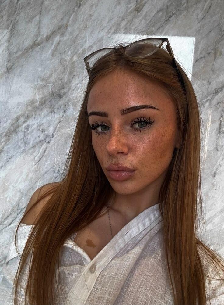 A portrait of a young woman with freckles, wearing a white off-shoulder shirt and sunglasses on her head, with a marble wall background.