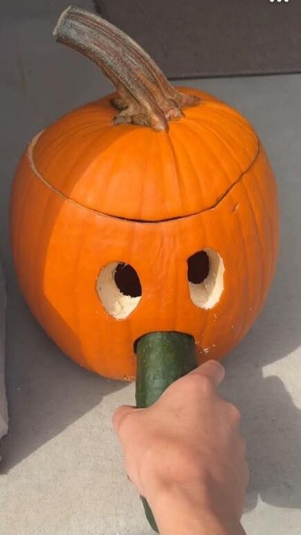 A carved pumpkin with two eye holes and a small green vegetable (looks like a cucumber or zucchini) being held up to its nose by a hand.