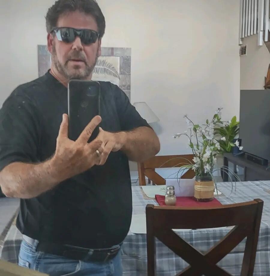 A man in sunglasses and a black t-shirt takes a selfie in a mirror, showing his reflection and part of a dining room.