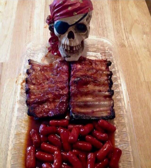 A skull decoration with an eye patch and red bandana sits atop a tray of cooked bacon strips with a bed of red sausages.