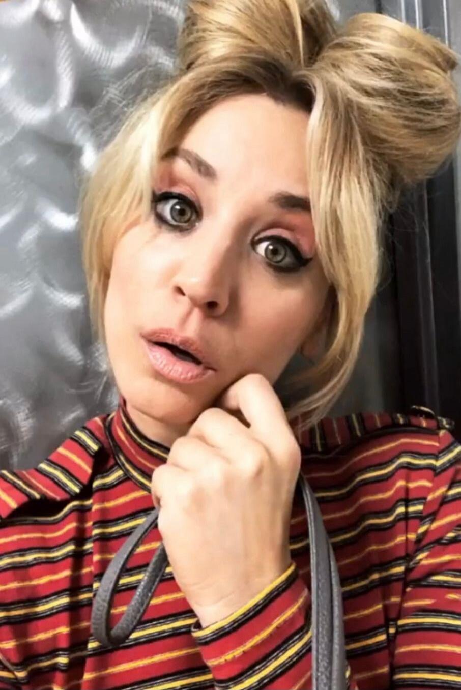 A close-up selfie of a blonde woman with her hair styled in two buns, wearing heavy eye makeup and a striped red, yellow, and black top. She has an expressive look with her mouth slightly open and one hand near her chin, holding a strap.