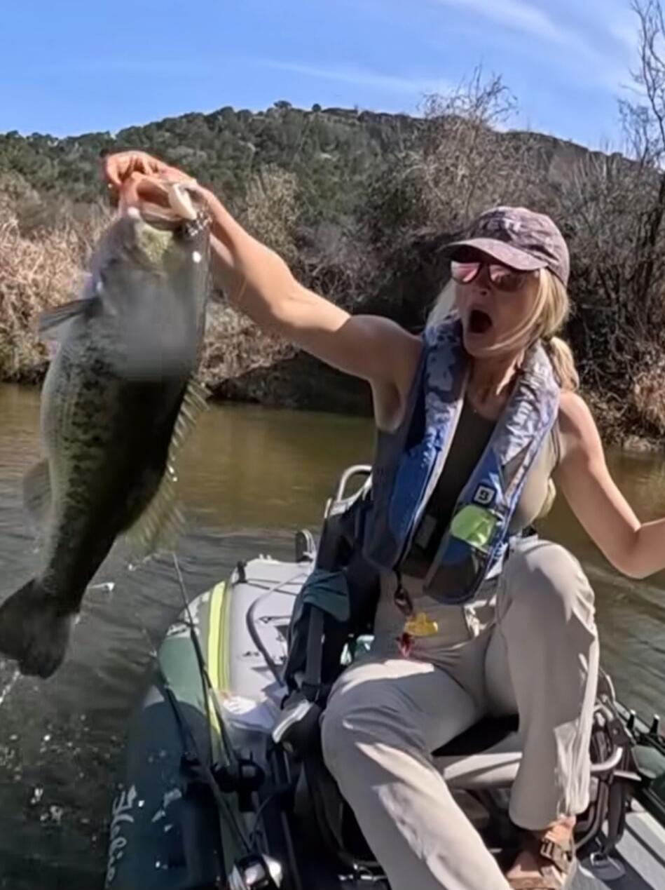 A woman in a kayak excitedly holds up a large fish she just caught, showing a surprised and joyful expression.