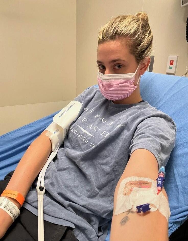 A woman in a hospital bed, wearing a pink and white striped mask, with an IV drip in her left arm and a blood pressure cuff on her right arm. She is wearing a blue t-shirt with 'ROSEMARY BEACH ESTABLISHED 19' visible. A hospital wristband on her right wrist shows 'LAC REPEAT LAC'.