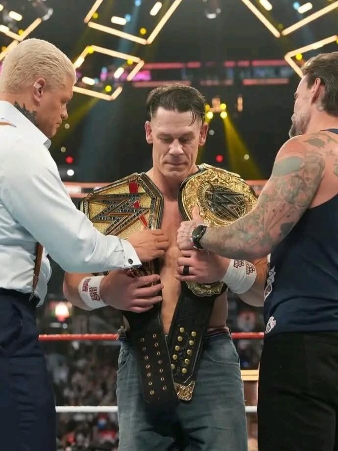 The image shows a wrestler with two championship belts being presented by two other men in a wrestling ring.
