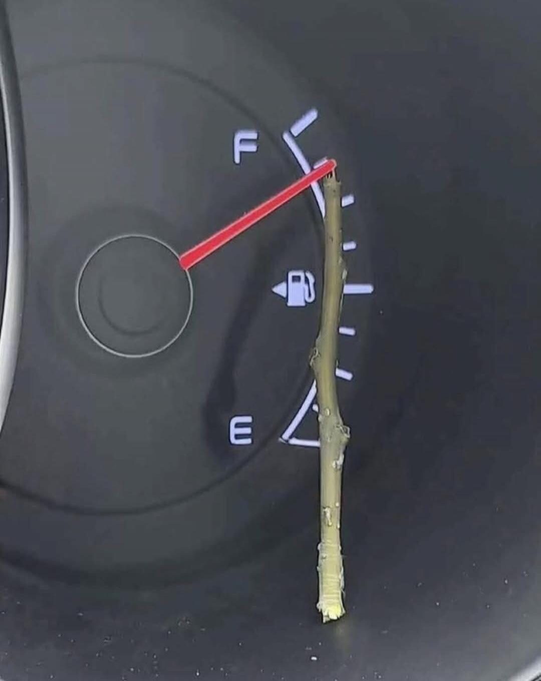 A car's fuel gauge shows the needle pointing to 'F' (Full), but a small stick is propped against it, humorously holding it in place. The letters 'F' and 'E' are visible, along with a fuel pump icon.