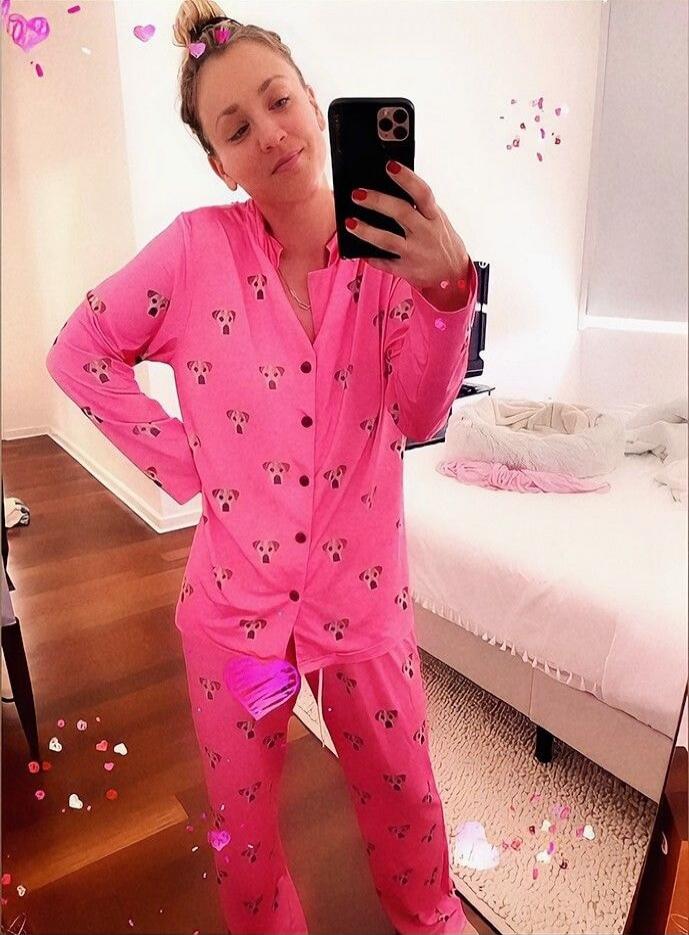 A woman in pink pajamas with a dog pattern takes a mirror selfie. Her hair is tied in a bun. There are heart-shaped decorations around her.