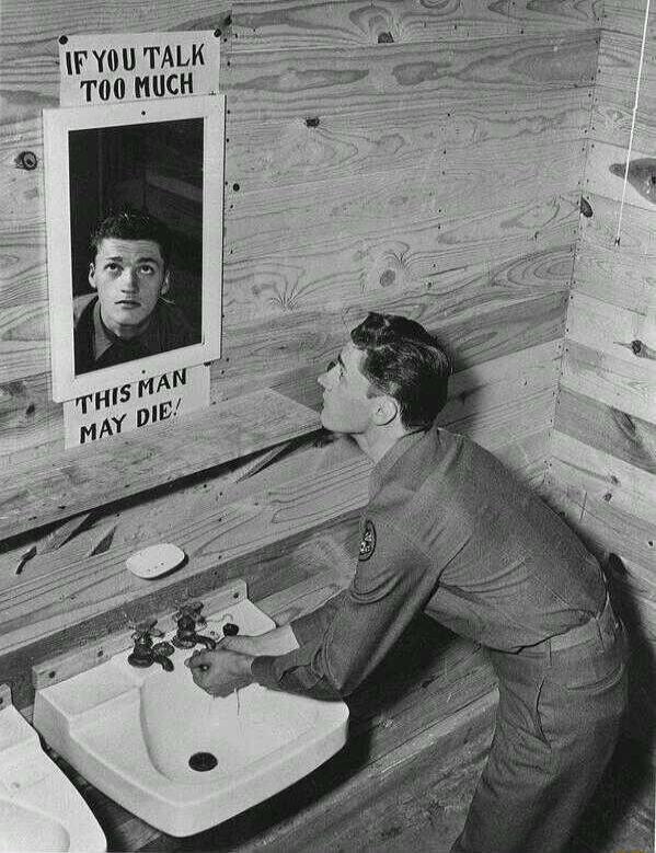 A man in uniform is washing his hands at a sink in a wooden room. Above the sink, a poster reads: 'IF YOU TALK TOO MUCH THIS MAN MAY DIE!' The poster features a black and white photo of another man looking upwards.