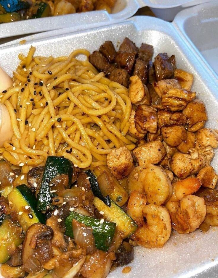 A delicious takeout meal featuring lo mein noodles topped with black sesame seeds, alongside grilled steak cubes, seasoned chicken pieces, and sautéed shrimp. The dish also includes stir-fried vegetables such as zucchini, mushrooms, and onions, all served in a white styrofoam container.