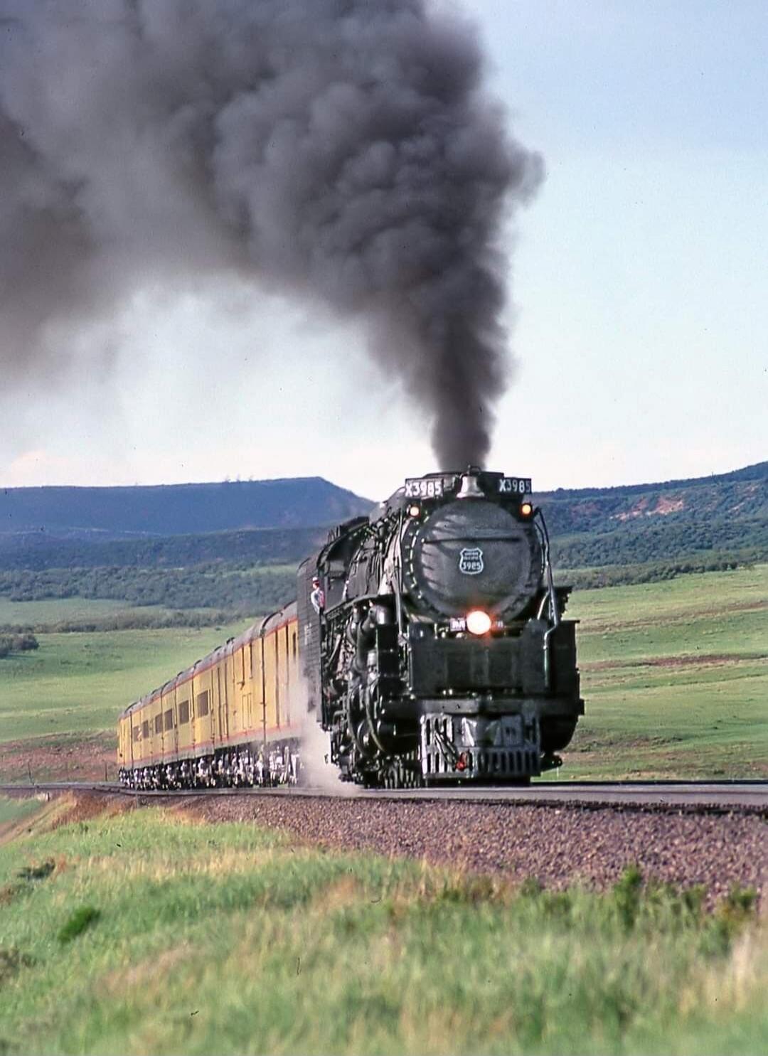 Steam locomotive pulling a long yellow passenger train through a rural landscape with a large black smoke plume.