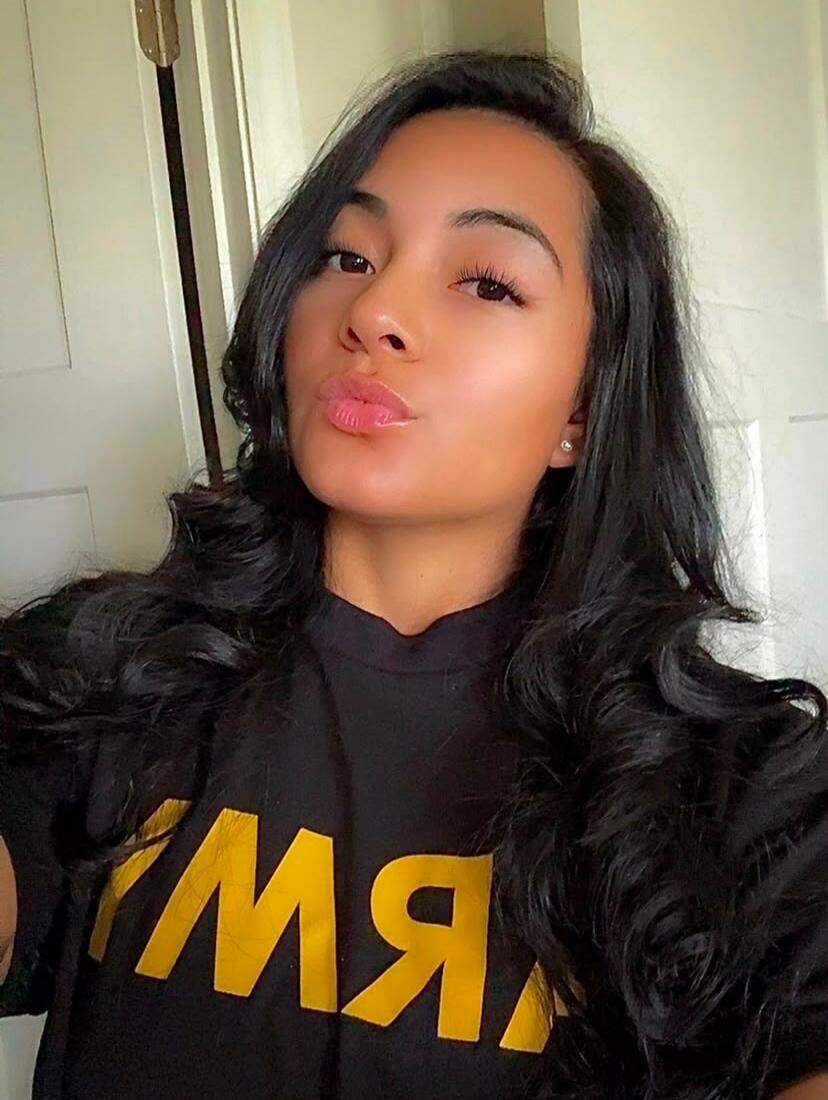 A woman with long dark hair, wearing a black t-shirt with yellow text, making a kissing face. The text on her shirt appears to be 'YMW' (mirrored).