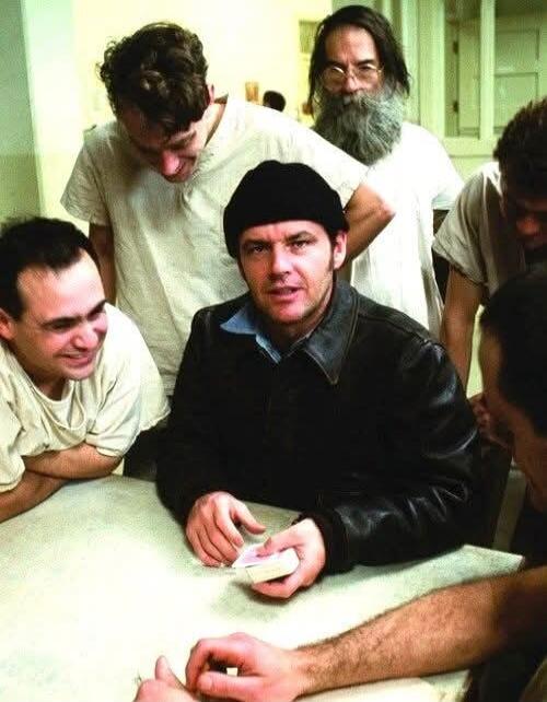A group of men, including Jack Nicholson as Randle McMurphy, are gathered around a table. McMurphy is holding a deck of cards, looking directly at the viewer. Other men are observing him, some smiling, in what appears to be a hospital or asylum setting.