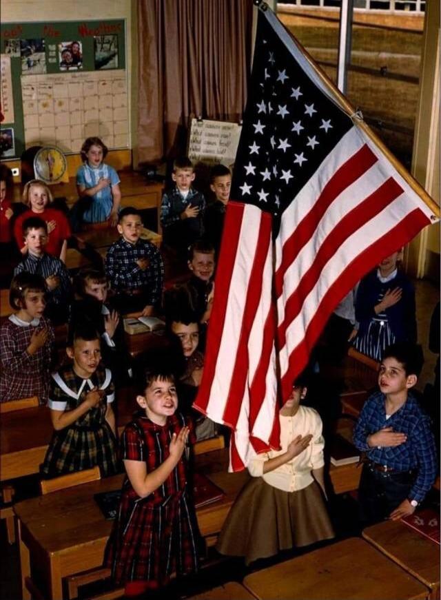 Children in a classroom with an American flag, hands over hearts, likely reciting the Pledge of Allegiance. Text on board: 'What comes next?', 'The Weather'.