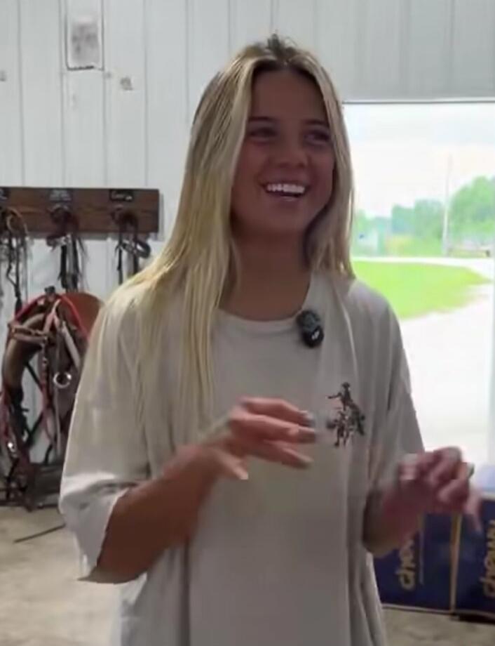 A young blonde woman is smiling and gesturing with her hands in what appears to be a stable or barn setting. Horse tack is visible in the background.