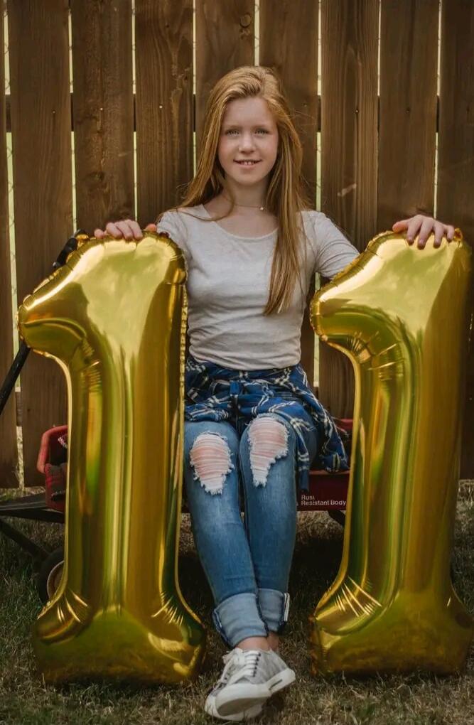 A young girl is sitting outdoors, holding two large golden balloons shaped like the number '1', forming '11'. She is smiling at the camera. Text visible: 'Rusi Resistant Body'.