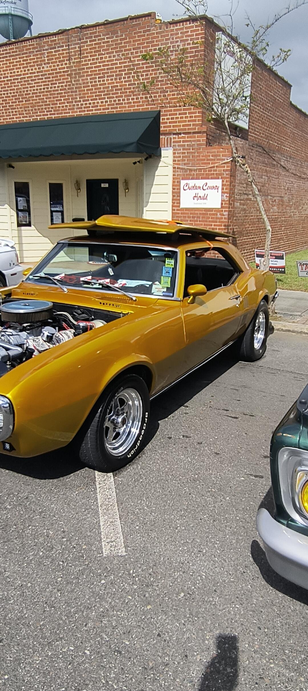 A golden classic muscle car, possibly a Chevrolet Camaro, is parked with its hood open, showcasing its engine. The car has chrome wheels with BFGoodrich tires. In the background, there's a brick building with a sign that reads 'Charlton County Herald'. Papers with '2019' and '2018' are visible inside the car.