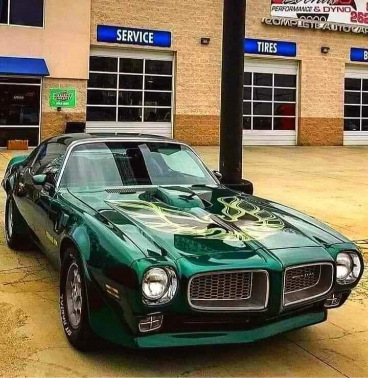 A classic green Pontiac Firebird Trans Am with a distinctive bird decal on the hood is parked in front of an auto service shop. Signs visible on the building include 