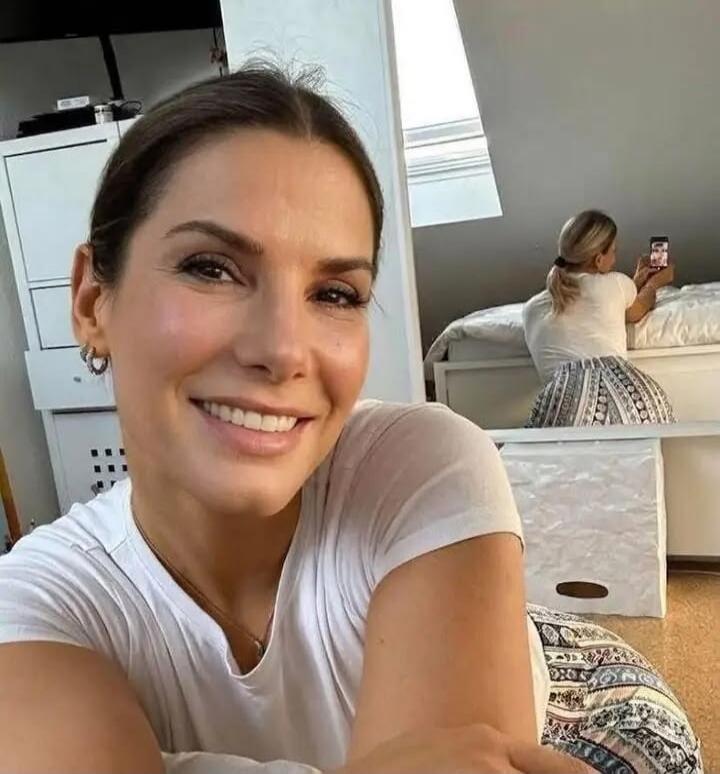 A woman taking a selfie, with another woman in the background also taking a photo with a phone, creating a layered perspective.