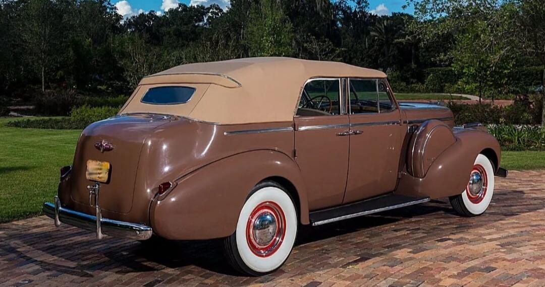 A vintage brown convertible car with a beige soft top, white wall tires, and chrome accents is parked on a brick driveway with green grass and trees in the background.
