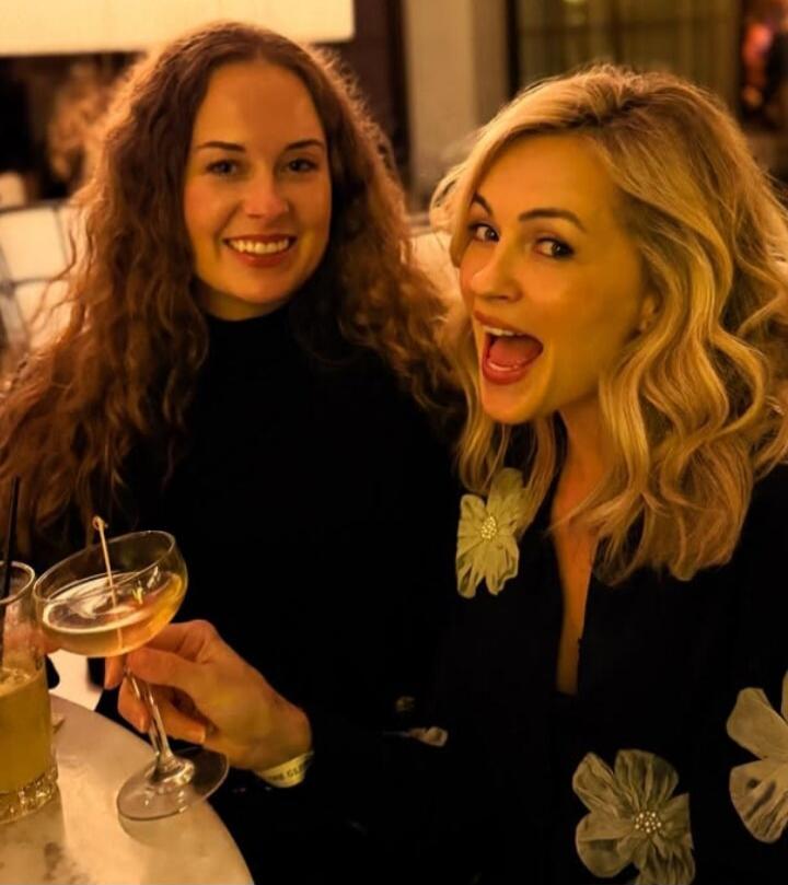 Two women are enjoying drinks at what appears to be a bar or restaurant. One woman with long curly brown hair is smiling, while the other with blonde wavy hair holds a cocktail and looks excited with an open mouth.